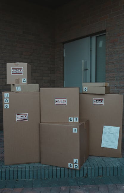 Several large cardboard boxes, some sealed with packing tape and marked with 'FRAGILE' labels and handling symbols, are stacked outside a building near a doorway. The boxes vary in size, with some placed on the ground and others positioned atop each other, indicating ongoing packing or loading processes typical of a home relocation. The boxes feature printed labels and some have white paper documents attached, possibly packing lists. The scene is set on a paved area with a brick building wall in the background and a metal door with a handle behind the boxes. This image reflects the logistical stage of furniture transport and packing involved in professional removals services, such as those provided by Man and Van South Lambeth, supporting smooth and careful home moving operations.