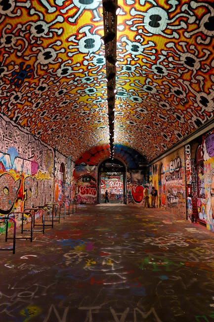 An underground passageway or tunnel covered with colorful graffiti on the walls and ceiling, featuring various tags, illustrations, and messages. The ceiling is decorated with a pattern of skull-like designs in orange, white, and black. The floor is also painted with graffiti, including various shapes and words. Along the sides, metal benches are positioned, and the area appears well-lit with artificial lighting. The tunnel is part of an urban environment, possibly used for recreational or artistic purposes, and the setting reflects a vibrant street art scene. This environment may be associated with cityscape elements relevant to house removals or moving services, such as transporting personal belongings through complex or artistic spaces, although the image primarily emphasizes the visual art scene.