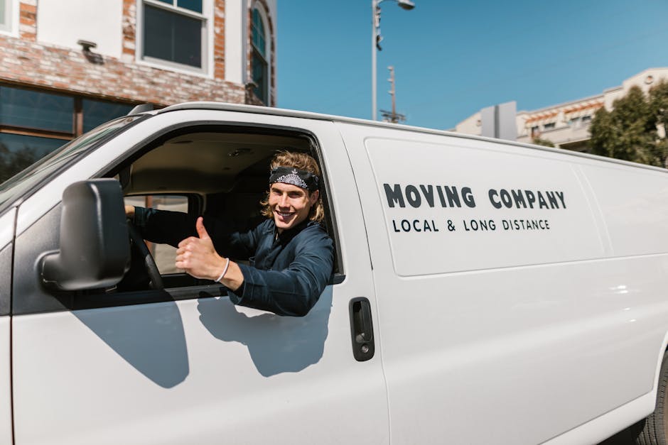 A young man with long, wavy hair wearing a black bandana and a dark long-sleeved shirt is sitting in the driver's seat of a white van belonging to Man and Van South Lambeth, a local removals company.
