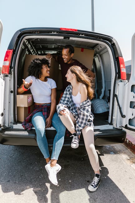 A young man with long, wavy hair wearing a black bandana and a dark long-sleeved shirt is sitting in the driver's seat of a white van belonging to Man and Van South Lambeth, a local removals company.