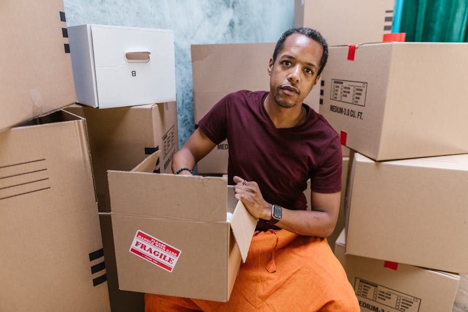 A man with dark skin and short hair, wearing a burgundy t-shirt and orange trousers, is seated amidst several cardboard moving boxes inside a property. He is holding and examining an open box with a focused expression, surrounded by additional boxes of varying sizes, some labeled with fragile stickers. The boxes are stacked and positioned closely around him, with some boxes on the floor and others placed on a surface. The background shows a partially visible doorway, and natural light illuminates the scene, highlighting the packing and loading process typical of home relocation services. This image exemplifies the packing phase of furniture transport and moving logistics undertaken by Man and Van South Lambeth, especially relevant for student flat removals in Brixton Lambeth, illustrating careful handling of household items during a house move.