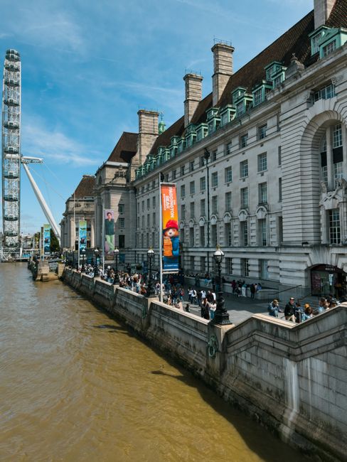 A large historic white building with multiple chimneys, situated alongside the River Thames in London, with pedestrians walking along the riverside pavement. Visible in front of the building are banners advertising a LEGO event, and a Ferris wheel can be seen to the left. The pavement appears to be used for loading and unloading, with some people carrying boxes or bags, possibly related to home relocation activities. Inside an open doorway near the building, a man is seen lifting or moving a box wrapped in protective packing materials, with additional cardboard boxes, plastic wrapping, and moving blankets visible on the ground nearby. A man-and-van service vehicle is parked close to the scene, indicating transportation support for furniture transport or packing and moving tasks associated with house removals. The environment is lively, daytime, with bright weather and clear skies, and the scene captures elements relevant to logistics involved in house removals and home relocation in a busy urban setting.