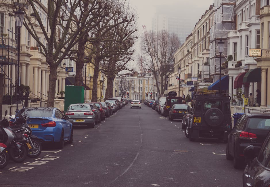 A residential street scene during daytime in South Lambeth, showing parked cars lining both sides of the road in front of Victorian-style terraced houses with white facades. Several vehicles, including a dark green Land Rover and a light grey van, are positioned near the pavement, some possibly involved in home relocation or furniture transport activities by Man and Van South Lambeth. The street is lined with leafless trees, and the sky appears overcast. The front gardens of the houses display decorative elements such as potted plants and awnings, indicating typical urban living spaces. No moving action is visible, but the arrangement suggests preparations for packing, loading, or unloading furniture and boxes as part of a packing and moving service, with the environment suitable for vehicle access and short-term parking during home removals.