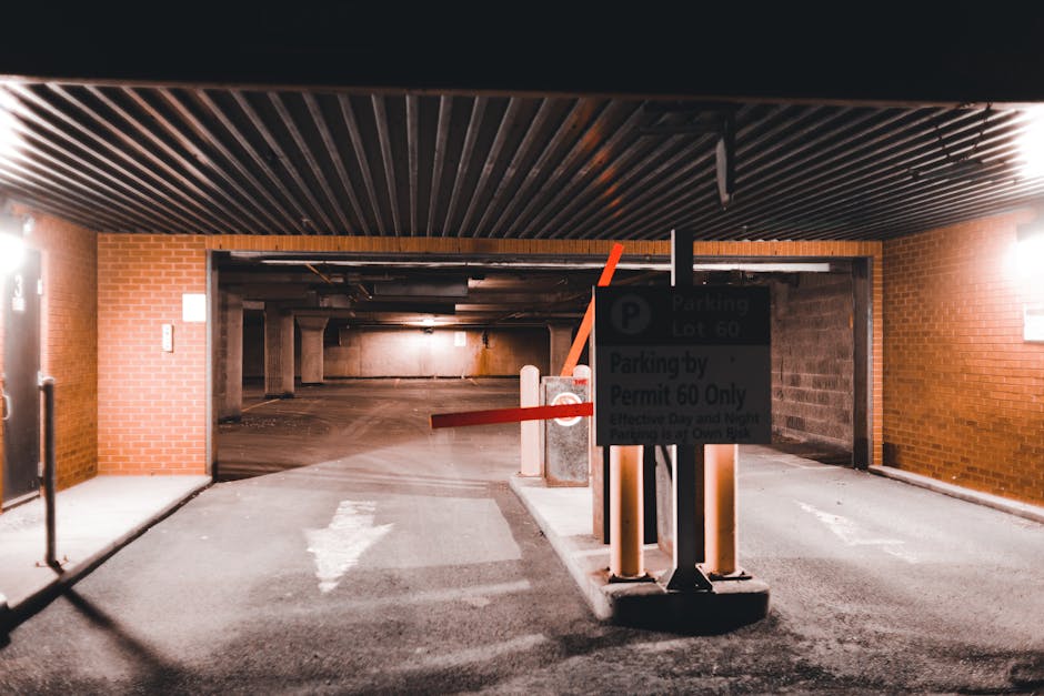 Inside a multi-story underground parking garage with a brick and concrete structure, an entrance to an indoor car park is partially visible with a black ceiling featuring parallel wooden slats and soft lighting. In the foreground, a parking control barrier with a red horizontal arm is lowered across a designated parking entry point. Nearby, a black and white parking regulation sign indicating 'Parking Lot 60' and 'Parking by Permit 60 Only' is mounted on a black post, partially obscuring the barrier. The ground is made of concrete with a white directional arrow painted on it, guiding vehicles forward. Behind the barrier, the parking area extends into a darker space with multiple concrete pillars supporting the ceiling and a few parked cars visible in the distance. The environment appears well-lit with artificial lighting, emphasizing the functionality of the parking system connected to urban house removals or furniture transport logistics, such as those carried out by Man and Van South Lambeth in coordination with local parking policies for residential relocations.
