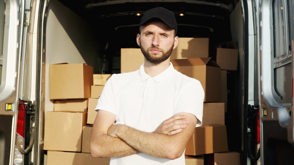A young man with a serious expression, wearing a black cap and a white polo shirt, stands with his arms crossed inside an open van loaded with cardboard moving boxes and plastic wrapping materials. The van, parked on a pavement outside a property, shows part of the vehicle's interior filled with packed items ready for home relocation or furniture transport. The boxes vary in size and are neatly stacked against the van's interior walls, with some wrapped in protective plastic to prevent damage during transit. The lighting appears natural, illuminating the packed items and the man, reflecting the typical loading process involved in professional removals services by Man and Van South Lambeth. This scene captures the key elements of packing, loading, and transportation involved in moving services for a residential move in the Kennington Oval area of South Lambeth.