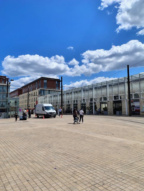 The image shows an outdoor urban setting with a large, open paved area in the foreground, made of beige bricks. Several people are visible, some walking and others standing, with a few pushing carts or carrying items. In the middle ground, there is a white delivery van parked near the building, which features a modern glass and metal facade with large windows and vertical structural supports. To the left, a small group of individuals appears to be engaged in a conversation or preparing for a home relocation or furniture transport process. The background includes a multi-storey building with a combination of brickwork and glass windows, and above, the sky is bright blue with scattered white clouds, suggesting clear weather suitable for moving or packing activities. The scene reflects typical elements involved in house removals or packing and moving services, with clear emphasis on the loading or unloading area outside a commercial or residential structure, as noted by Man and Van South Lambeth.