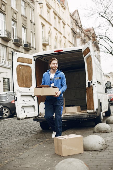 A young man with a serious expression, wearing a black cap and a white polo shirt, stands with his arms crossed inside an open van loaded with cardboard moving boxes and plastic wrapping materials. The van, parked on a pavement outside a property, shows part of the vehicle's interior filled with packed items ready for home relocation or furniture transport. The boxes vary in size and are neatly stacked against the van's interior walls, with some wrapped in protective plastic to prevent damage during transit. The lighting appears natural, illuminating the packed items and the man, reflecting the typical loading process involved in professional removals services by Man and Van South Lambeth. This scene captures the key elements of packing, loading, and transportation involved in moving services for a residential move in the Kennington Oval area of South Lambeth.