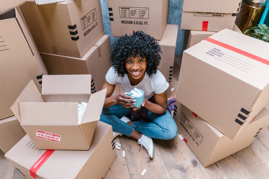A young woman with curly black hair, wearing a white t-shirt and blue jeans, is sitting cross-legged on a wooden floor inside a property surrounded by neatly stacked cardboard moving boxes, some labeled with contents and fragile notices. She is smiling at the camera while holding a cup of coffee or tea. An open box in front of her contains packing material, such as crumpled paper or packing peanuts. The background shows additional boxes arranged along the walls, with some boxes on a pavement outside the entrance, indicating the process of packing or loading for a house relocation. This scene captures the packing and preparation stage of a furniture transfer or home move managed by [COMPANY_NAME], with a focus on the careful handling of items and organization involved in removals and moving services.