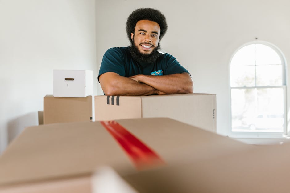 A young man with a dark beard and curly hair, smiling and standing behind a stack of brown cardboard moving boxes inside a well-lit room with a large arched window in the background. The boxes vary in size, some sealed with packing tape and one featuring a red stripe, indicating they are prepared for a house removal or home relocation. The environment appears clean and minimalistic, with the boxes positioned on a flat surface, possibly a table or floor, as part of the packing process organized by Man and Van South Lambeth. This scene illustrates a typical packing and loading process involved in furniture transport and moving services, highlighting the company's role in facilitating smooth removals for residential clients while ensuring secure and efficient handling of household items.