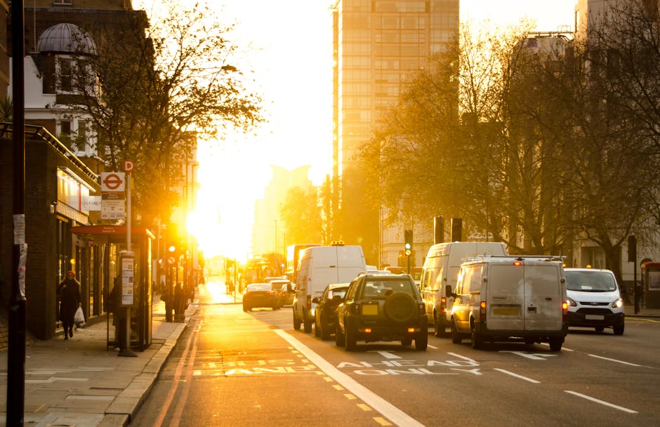 A busy city street during sunset in South Lambeth with a row of parked and moving vehicles, including white vans, small cars, and a white truck, along the curbside. The pavement features a bus stop shelter with a timetable, and a few pedestrians walking along the sidewalk. Tall buildings and leafless trees line both sides of the street, with the sunlight casting a warm glow over the scene. The vehicles are positioned in the loading area, likely involved in a home relocation or furniture transport process managed by Man and Van South Lambeth, as part of their removals services. The scene captures the typical outdoor environment seen during packing, loading, or unloading activities in the context of same-day removals and moving logistics.