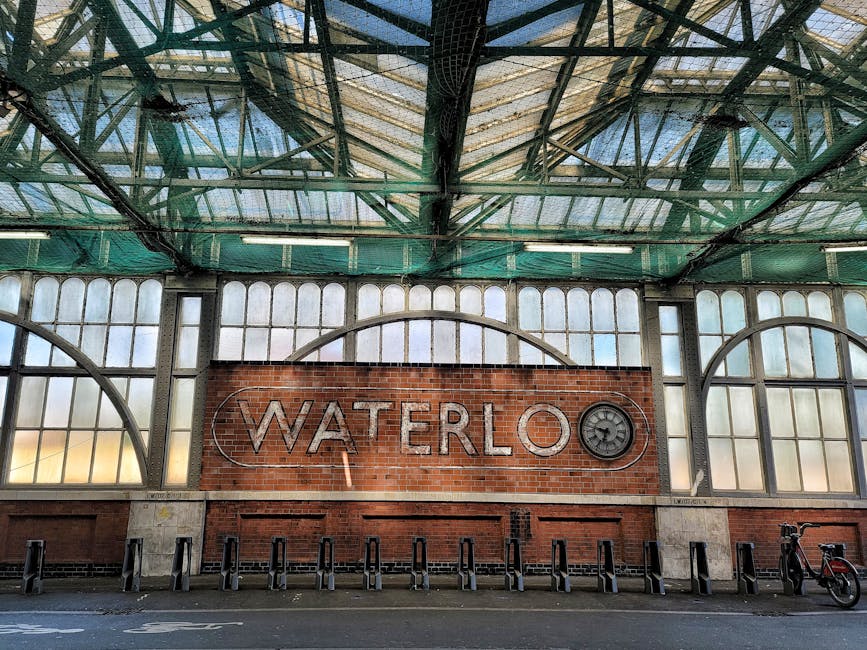 Inside Waterloo station, a large brick wall features illuminated metal letters spelling 'WATERLOO' with a clock symbol embedded in the letter 'O'. Above, the station's high, arched glass roof is supported by metal framework, allowing natural light to filter through. On the pavement in front of the wall, there are several black bike racks and a single red bicycle parked to the right. The scene captures the station's architectural details and spacious interior, which may be part of a home relocation or moving logistics process involving furniture transport and packing, as managed by Man and Van South Lambeth, specializing in removals.
