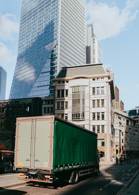 A large green delivery truck with a white rear door is parked on a city street in front of modern high-rise buildings and older stone structures, during daylight hours. The truck is positioned adjacent to the curb, possibly waiting for loading or unloading activities as part of a home relocation process. The street scene includes some pedestrians and cyclists, with the surrounding architecture featuring glass facades and older architectural styles typical of central London. The weather appears clear with a blue sky and a few clouds, providing good natural lighting, which highlights the building facades and the loading area of the van. As part of moving and packing services by Man and Van South Lambeth, the truck is ready for furniture transport and equipment for packing and moving household items in preparation for a house removal near Lambeth North station and Waterloo.