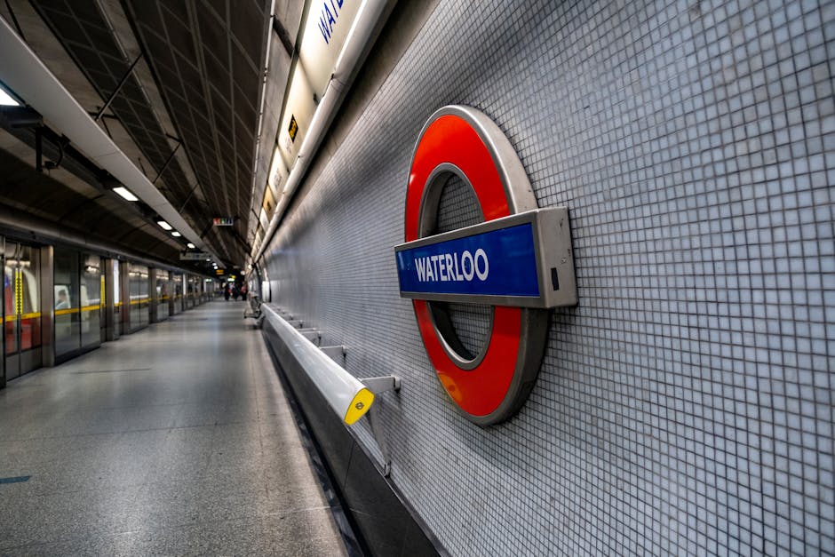 Inside Waterloo station, a large brick wall features illuminated metal letters spelling 'WATERLOO' with a clock symbol embedded in the letter 'O'. Above, the station's high, arched glass roof is supported by metal framework, allowing natural light to filter through. On the pavement in front of the wall, there are several black bike racks and a single red bicycle parked to the right. The scene captures the station's architectural details and spacious interior, which may be part of a home relocation or moving logistics process involving furniture transport and packing, as managed by Man and Van South Lambeth, specializing in removals.