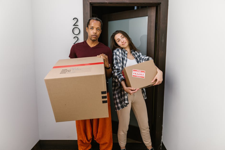 A man and a woman stand inside a doorway of a residential property, each holding a cardboard box used for house relocation. The man, dressed in a maroon T-shirt and orange trousers, holds a larger box with a label indicating fragile contents. The woman, wearing a plaid shirt over a white T-shirt and beige trousers, holds a smaller box with a red and white label also indicating fragility. The scene occurs near a doorframe, with a dark wooden door and the property number '2002' visible on the wall beside them. The background includes indoor wall surfaces painted in light color; the floor is dark, contrasting with the white walls. The overall setting suggests packing and loading processes involved in furniture transport and the house removal service provided by Man and Van South Lambeth, supporting home relocation tasks and moving logistics.
