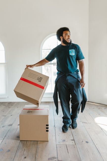 A man with dark curly hair and a beard is inside a bright, empty room with white walls and a large arched window allowing natural light to fill the space. He is dressed in casual work clothing, including a blue t-shirt with a logo on the chest and dark pants, with a jacket tied around his waist. He is carefully lifting a cardboard box wrapped with red packing tape, preparing to load or carry it for home relocation or furniture transport. There are two other cardboard boxes on the wooden floor, one on top of the other, both sealed with tape. The scene depicts a loading process associated with house removals and packing and moving services, emphasizing careful handling of packed belongings. Man and Van South Lambeth, a removals specialist company, appears to be involved in this professional moving activity, with the focus on efficient and secure transition of household items during a house move or furniture transport.