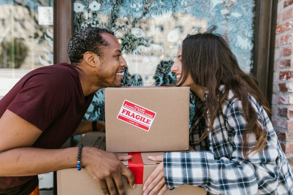 A man and a woman stand inside a doorway of a residential property, each holding a cardboard box used for house relocation. The man, dressed in a maroon T-shirt and orange trousers, holds a larger box with a label indicating fragile contents. The woman, wearing a plaid shirt over a white T-shirt and beige trousers, holds a smaller box with a red and white label also indicating fragility. The scene occurs near a doorframe, with a dark wooden door and the property number '2002' visible on the wall beside them. The background includes indoor wall surfaces painted in light color; the floor is dark, contrasting with the white walls. The overall setting suggests packing and loading processes involved in furniture transport and the house removal service provided by Man and Van South Lambeth, supporting home relocation tasks and moving logistics.